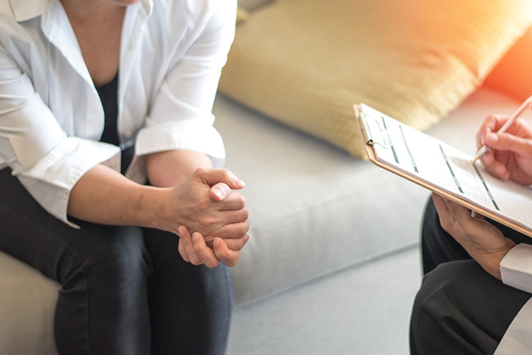 A close up of two people sitting. One has a clipboard and the other is clasping their hands